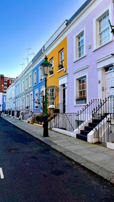 A row of colorful terraced houses on a street in West Kensington, featuring pastel facades in shades of blue, yellow, and lavender, with white-framed windows and black wrought iron fences along the si