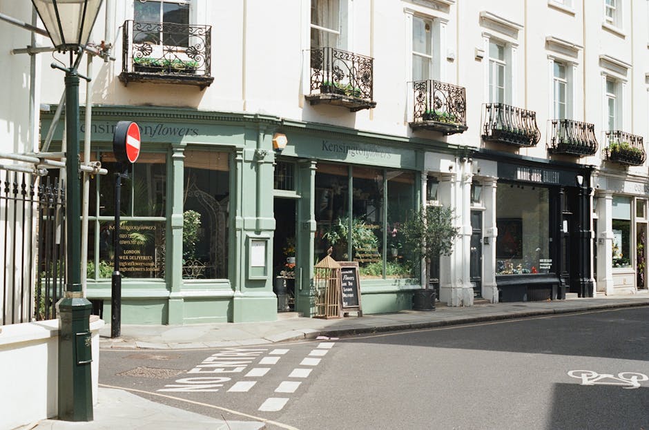 Exterior view of a narrow street in West Kensington featuring a group of ground-floor shops with large glass windows, including a flower shop painted in pastel green with floral displays visible inside, and a gallery with a black façade. The shops are part of a classic-style terraced building with white walls and black wrought iron balconies on the upper floors. A small white chalkboard sign is positioned on the sidewalk outside the flower shop, possibly displaying promotional or informational notices. In front of the shops, a sidewalk runs along the street, and a bicycle lane is marked with white lines on the asphalt. A metal bollard and a temporary pole are situated near the curb, and a street sign indicating a no-entry restriction is mounted on a pole nearby. The overall scene depicts a typical urban setting appropriate for home relocations or moving services, with elements such as the storefronts, street markings, and urban environment in clear view, suitable for illustrating house removals or furniture transport concepts. The lighting suggests daytime with natural sunlight illuminating the scene.