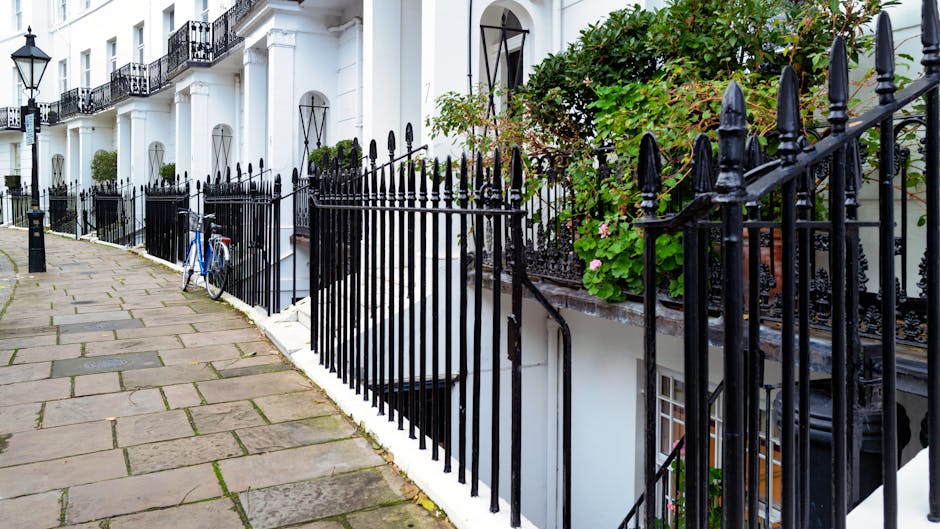 A row of white terraced houses with black wrought iron railings along a paved sidewalk on a sunny day. A blue bicycle is parked against one of the railings near a downward staircase leading to an underground entrance. Green plants and pink flowers grow in pots on the steps and along the front of the houses. In the foreground, the pavement is made of large, rectangular stone slabs, with a black bollard visible on the left. The area appears quiet, with no pedestrians present, and the scene captures a typical residential street suitable for home relocation and furniture transport services.