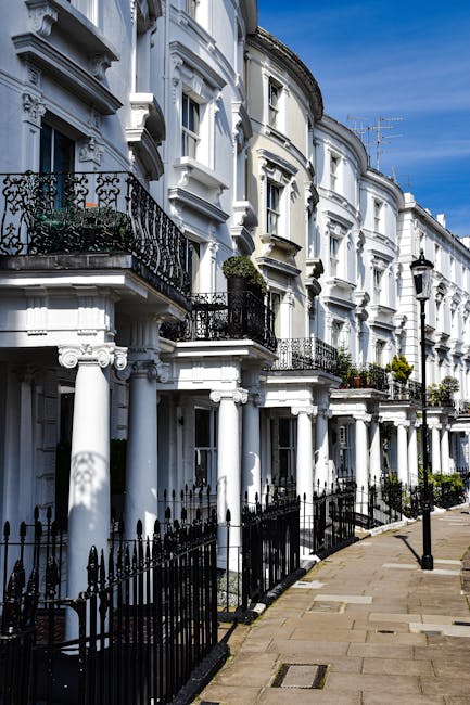 A row of white Victorian-style terraced houses on a residential street, featuring ornate cornices, decorative railings, and small balconies with black wrought iron railings. The buildings have large sash windows, some with flower boxes or potted plants, and are supported by white columns at ground level. The pavement in front is paved with stone slabs, with a black metal fence running along the edge. The scene is illuminated by daylight, with a clear blue sky and a few street lamps visible along the sidewalk. This setting reflects a typical urban neighborhood suitable for house removals and relocation services, with potential for loading and moving activities involving furniture and packing materials inside the properties.