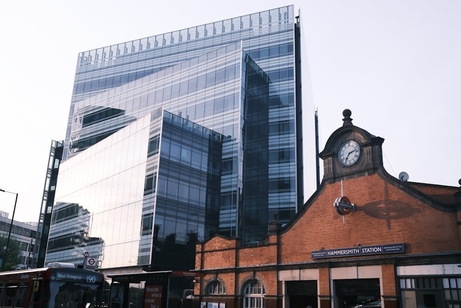 A modern glass office building with reflective windows showing the sky and surrounding structures, located behind Hammersmith Station in West Kensington. In the foreground, the historic brick facade and clock of Hammersmith Station are visible, with a black and white sign indicating the station's name. The image captures the area where furniture and large packing boxes could be prepared for a professional removal or moving service, with the reflective glass indicating recent or ongoing home relocation or furniture transport activities close to the station area. The scene is well-lit with natural daylight, highlighting the contrast between the contemporary commercial architecture and the traditional station building, relevant to relocation logistics supported by [COMPANY_NAME].
