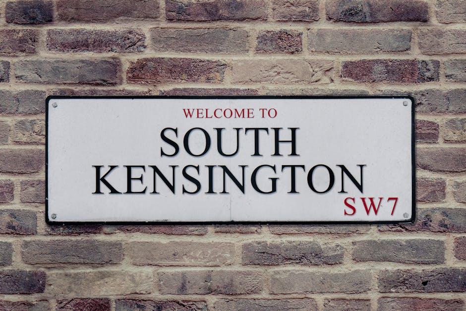 Close-up view of a rectangular street sign mounted on a brick wall, displaying the words 'WELCOME TO SOUTH KENSINGTON' in bold black letters with a red 'SW7' postcode in the bottom right corner. The sign is white with black border accents and is attached with four screws, one in each corner. The brick wall behind the sign consists of reddish-brown bricks with light mortar, providing a textured background. This setting is indicative of a typical residential or commercial property exterior in West Kensington, relevant to house and office removals, logistics, and packing processes by [COMPANY_NAME], supporting the overall theme of moving and furniture transport services in the West Kensington area.