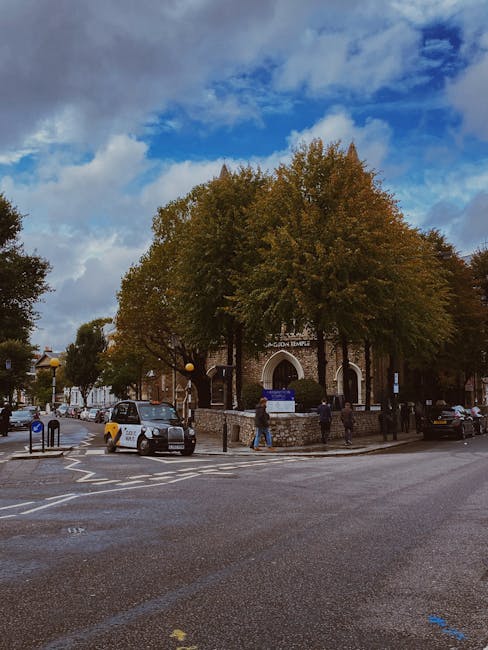 A blue bicycle with a black front basket is secured and leaning against a black wrought iron fence outside a residential property in West Kensington, with the front wheel touching the pavement. The si
