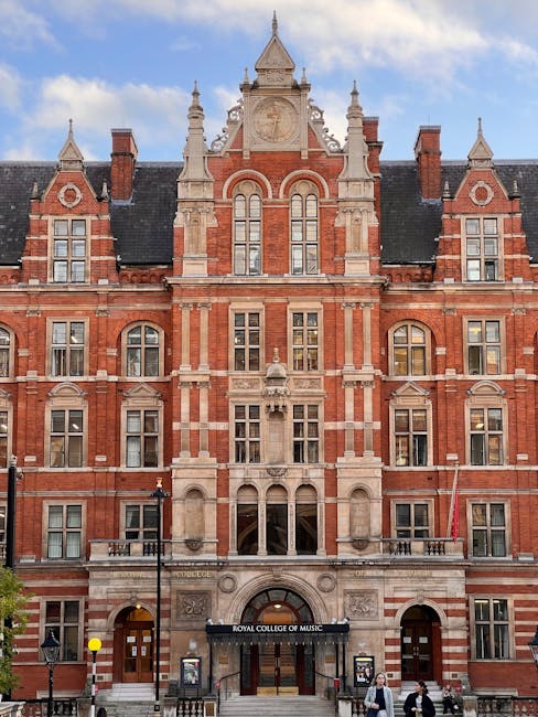 A residential street lined with parked cars on both sides, featuring a row of cherry blossom trees in full bloom with pink flowers and dark branches, extending over the pavement and road. The street is bordered by traditional terraced houses with white facades and large windows. The sky is partly cloudy with patches of blue visible. This scene illustrates a quiet neighbourhood during spring, suitable for house removals or relocation services, with no moving activities visible in the image. The image is captured during daytime, emphasizing the vibrant seasonal bloom and the typical urban streetscape associated with West Kensington, supporting the context of home relocation and furniture transport by removals specialists like those at removalcompanywestkensington.co.uk.