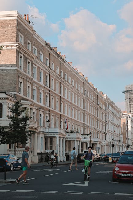 A row of multi-storey residential buildings with ornate architectural details, large windows, and white decorative trim, located along a busy street in West Kensington. In the foreground, pedestrians are crossing the road, including a person riding a bicycle and others walking, while parked cars line the pavement. The buildings feature brick and stone facades, with some windows partially open, and a clear blue sky overhead. This urban scene exemplifies typical London residential architecture and bustling city life, relevant to house removals and property relocation services offered by company. The image captures a moment during daytime, with natural light illuminating the scene, illustrating the environment in which home relocation and furniture transport services are often performed.