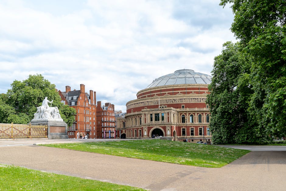 Image of a wide, outdoor view of the Royal Albert Hall in London, with its distinctive circular brick building and domed roof clearly visible. In the foreground, a paved pathway intersects a grassy area, leading towards the hall. To the left, there is a white marble statue on a pedestal, surrounded by a decorative metal fence. Large green trees border the scene on the right and left sides, providing shade and framing the building. The sky above is partly cloudy, with patches of blue visible. The setting appears calm, typical of a public park or garden area adjacent to the venue. This scene is associated with home relocation or moving services in the West Kensington area, highlighting the importance of careful furniture transport and packing during house removals, as sometimes seen in professional moving operations by companies like [COMPANY_NAME].