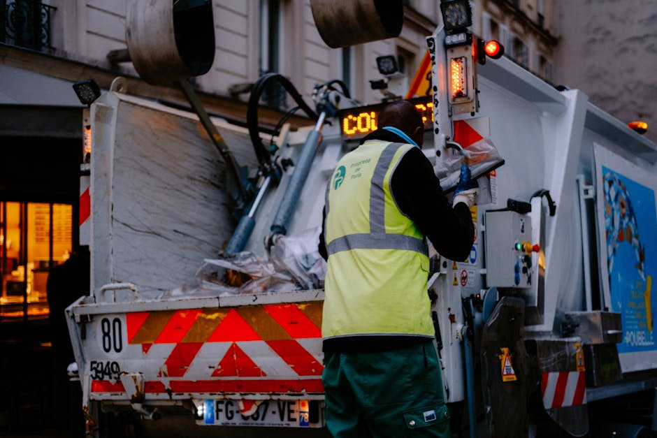 A person wearing a white shirt and beige trousers, with a red cardigan, is placing a clear plastic bottle into a grey recycling bin labelled 'PLASTIC'. The scene appears to be inside a home, near a ki
