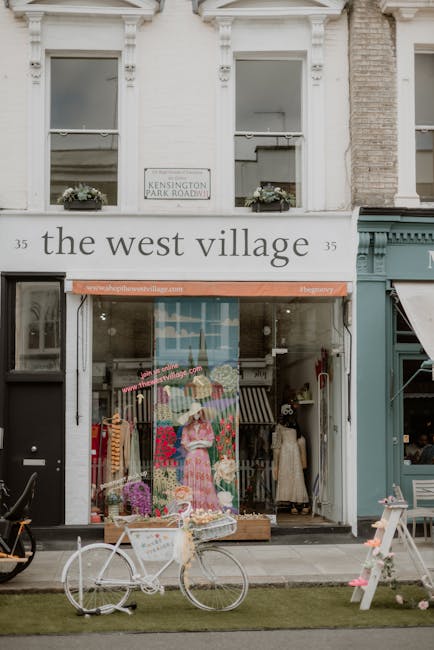 The image shows the exterior of a small retail shop named 'the west village' located at number 35, situated on a street in West Kensington. The shop's large front window displays colorful clothing and